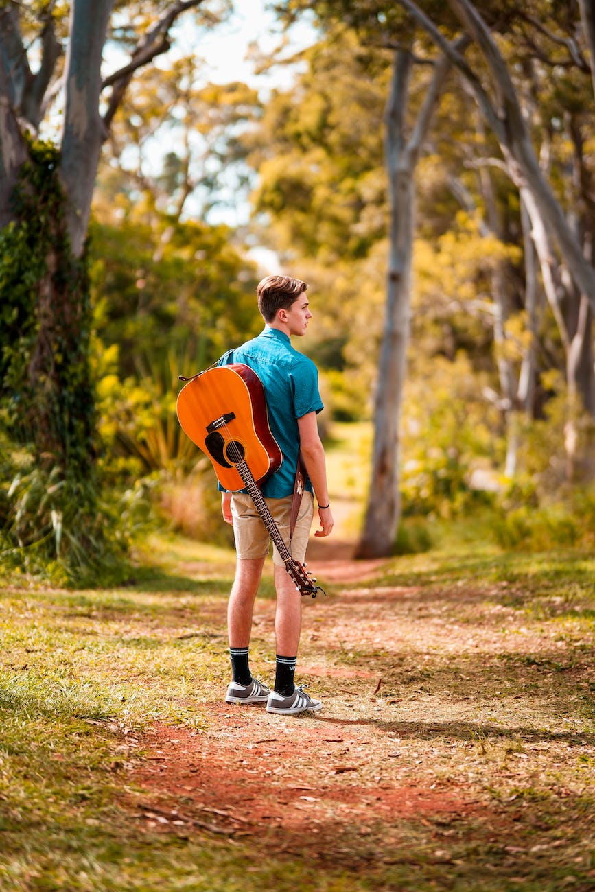 back view photo of man acoustic carrying guitar standing alone on trail looking to his right